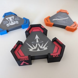 May include: Set of three triangular coasters with a gaming theme. Each coaster features a unique design with gray, black, red, blue, and orange accents. The coasters are stacked, showcasing their layered construction and detailed graphics.