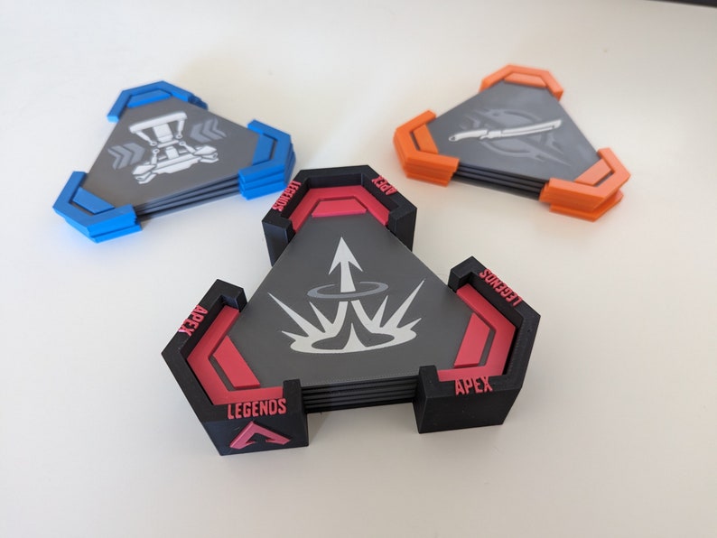May include: Set of three triangular coasters with a gaming theme. Each coaster features a unique design with gray, black, red, blue, and orange accents. The coasters are stacked, showcasing their layered construction and detailed graphics.