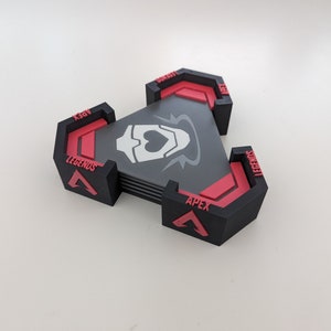May include: A stack of black and red triangular coasters with the text "APEX LEGENDS" and a white graphic. The coasters are arranged in a triangular shape. The background is a plain white surface.
