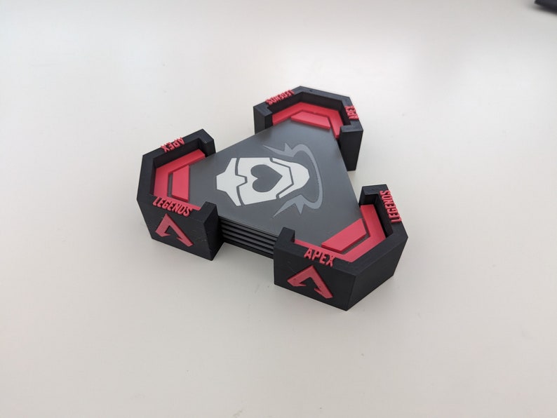 May include: A stack of black and red triangular coasters with the text "APEX LEGENDS" and a white graphic. The coasters are arranged in a triangular shape. The background is a plain white surface.