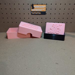 Könnte beinhalten: Zwei rosa Plastikboxen mit dem Aufdruck "Fight Club" auf der Oberseite. Die Boxen sind geöffnet und die linke Box hat einen Deckel.