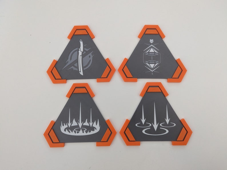 May include: Four gray, triangular game pieces with orange accents. Each piece features a unique white graphic: a knife, a geometric design, a flame-like shape, and arrows. The pieces are arranged on a white surface.