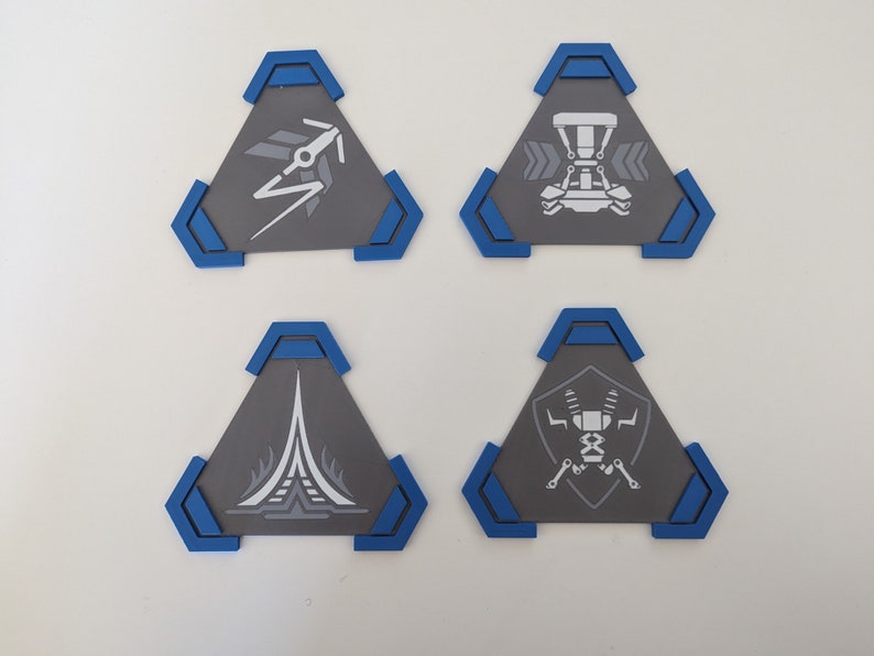 May include: Four gray, triangular coasters with blue trim. Each coaster features a different white graphic design: a stylized arrow, a vehicle, a tower, and a robot. These coasters are ideal for protecting surfaces from drink condensation.