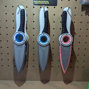 May include: Three 3D printed knives with different coloured handles. The knives are grey with white and black handles. The handles have a ring with a blue, blue, and red accent. The knives are hanging on a pegboard.