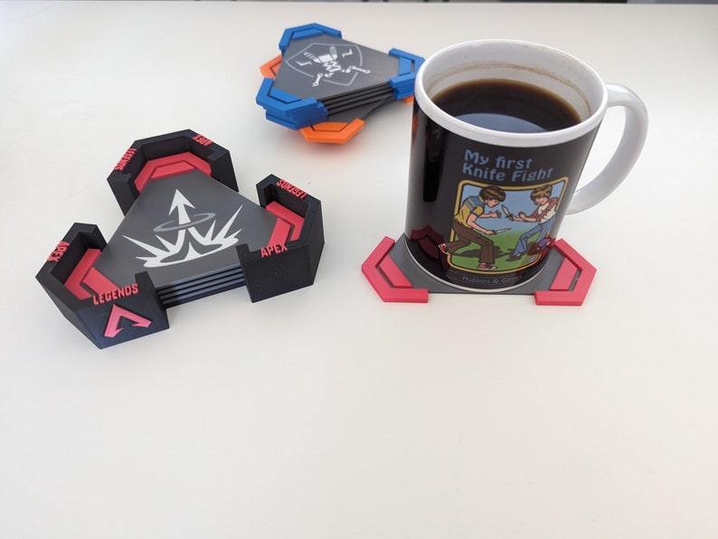 May include: A set of black, red, and gray coasters with the text "APEX LEGENDS" and a graphic design. A white mug with the text "My first Knife Fight" and a cartoon image sits on a matching coaster. Additional coasters are visible.