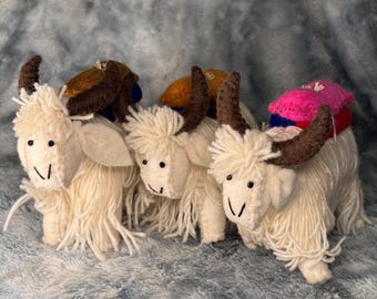 May include: Three white fluffy felt yak ornaments with brown horns and colorful felt saddle blankets. The yaks are standing in a row on a gray fuzzy surface.