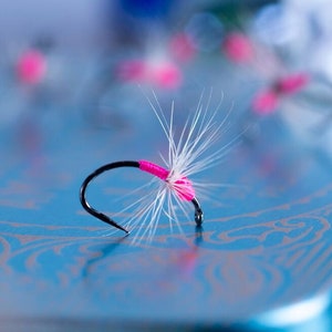 May include: Close-up of a black fishing hook with a bright pink band and white feather-like fibers. The hook is set against a blurred blue background with other similar hooks, suggesting a fishing theme.