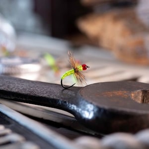 May include: A small green and red fly fishing lure with feathers and a hook, resting on a black metal wrench.
