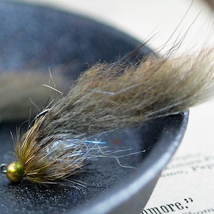 May include: Close-up of a handmade fishing fly. The fly features a gold-colored bead head, brown and tan fur, and iridescent accents. It is resting on a dark surface, possibly a fishing tackle box or tray.