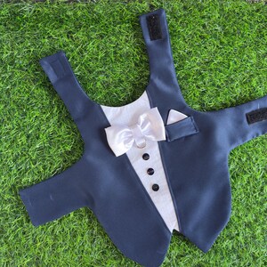 May include: A black tuxedo-style dog vest with a white bow tie and a pocket on the right side. The vest has a hook and loop closure on the back.