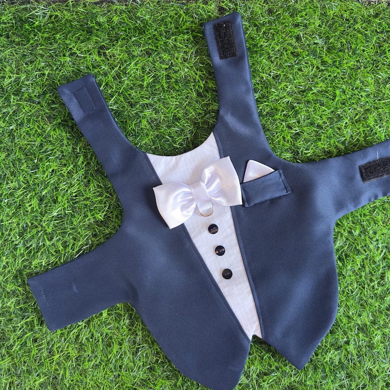 May include: A black tuxedo-style dog vest with a white bow tie and a pocket on the right side. The vest has a hook and loop closure on the back.