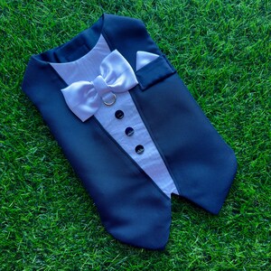 May include: A blue tuxedo-style dog vest with a white bow tie and black buttons. The vest is lying on a green grassy surface.