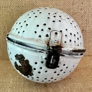 May include: A white, spherical, vintage kitchen strainer with numerous small holes. The strainer is made of metal and has a hinged lid with a black clasp. The enamel is chipped in places, revealing the metal underneath.