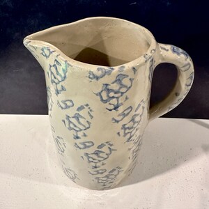 Op de afbeelding: Een witte keramische kan met een blauw en wit bloemenpatroon. De kan heeft een handvat en een tuit.