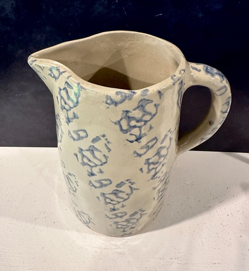 Op de afbeelding: Een witte keramische kan met een blauw en wit bloemenpatroon. De kan heeft een handvat en een tuit.