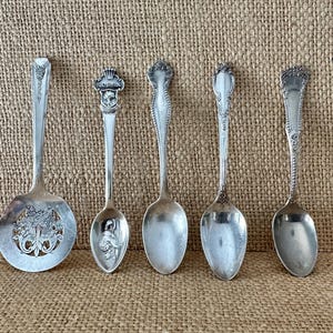 May include: Five antique silver spoons of varying designs are displayed. The spoons have ornate handles and bowls, with one featuring a pierced floral design. The spoons are arranged on a textured, neutral-toned surface.