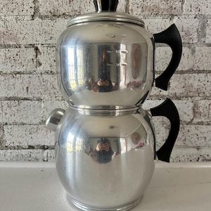 May include: A silver, two-tiered coffee maker with black handles. The top tier has a lid and a measurement scale on the side. The bottom tier has a spout for pouring coffee.