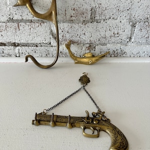 May include: Three brass wall hooks in the shape of a bird, a fish, and a pistol. The pistol hook has a chain and a decorative medallion.