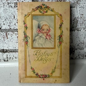 May include: Vintage book titled "Babys Days" with a baby portrait on the cover. The cover features a framed image of a baby in a white bonnet and dress, surrounded by floral and ribbon accents. The book has a cream-colored background.