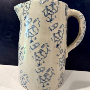 Op de afbeelding: Een witte keramische kan met een blauw-wit bloemenpatroon. De kan heeft een handvat en een tuit.