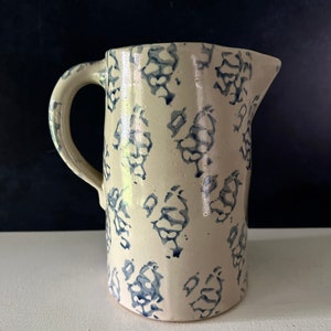 Op de afbeelding: Een witte keramische kan met een blauw en wit bloemenpatroon. De kan heeft een handvat en een brede opening.