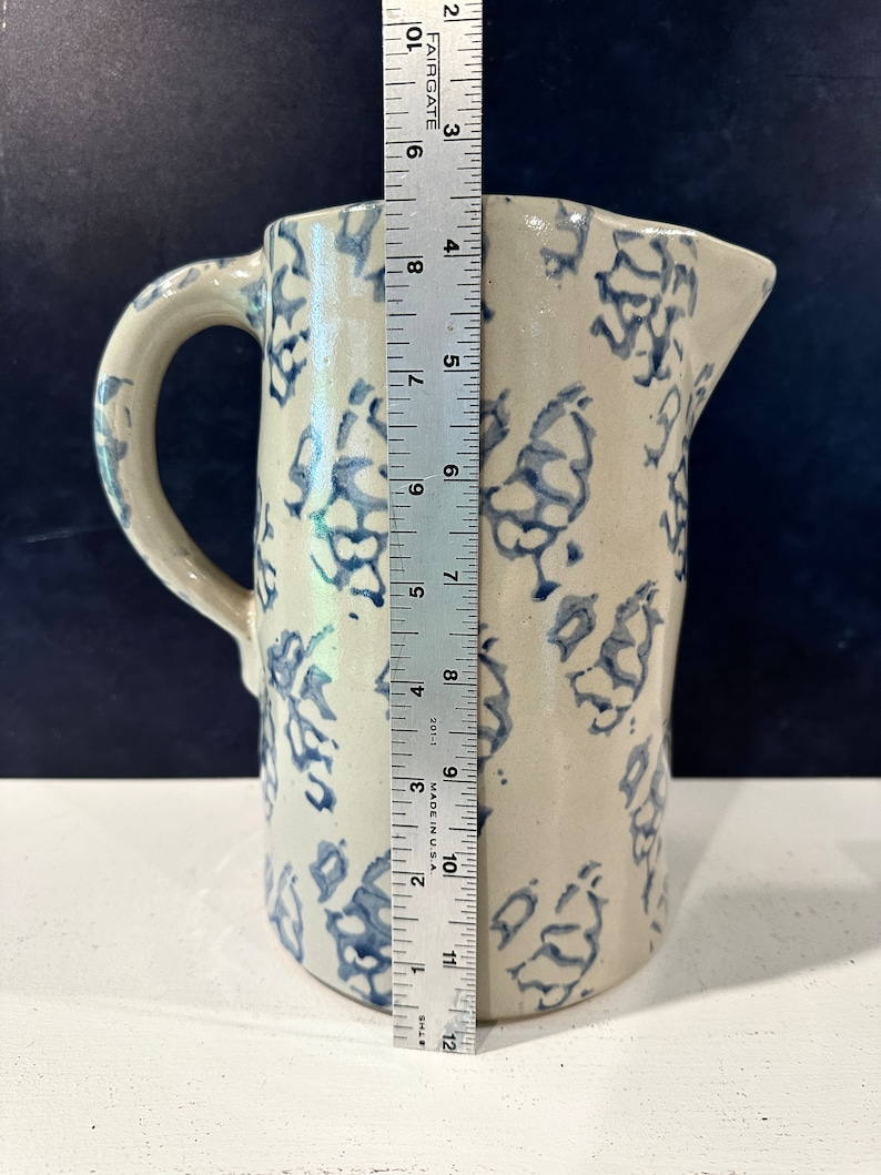 Op de afbeelding: Een witte keramische kan met een blauw-wit bloemenpatroon. De kan heeft een handvat en is ongeveer 30 cm hoog.