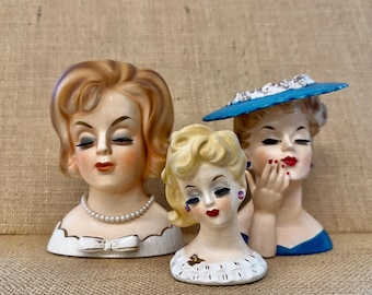 Peut inclure: Trois vases en céramique en forme de tête avec des visages aux traits peints. Le vase de gauche a des cheveux blonds et un collier de perles. Le vase central est plus petit avec des cheveux blonds et une fleur. Le vase de droite porte un chapeau bleu.