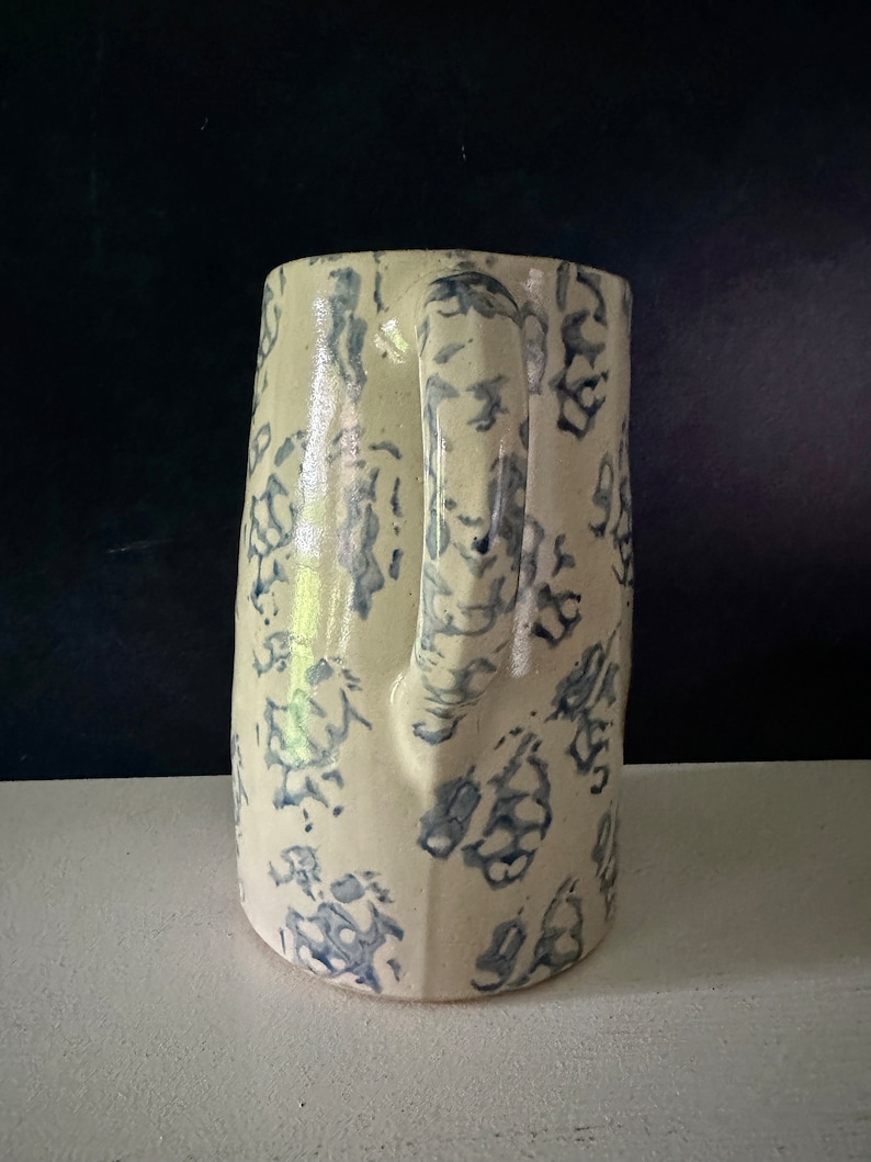 Op de afbeelding: Een witte keramische kan met een blauw bloemenpatroon. De kan heeft een handvat en een brede opening.