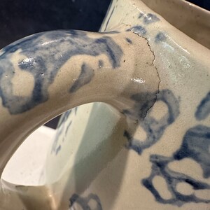 Op de afbeelding: Een witte keramische kan met een blauw bloemenmotief en een scheur in de handgreep.
