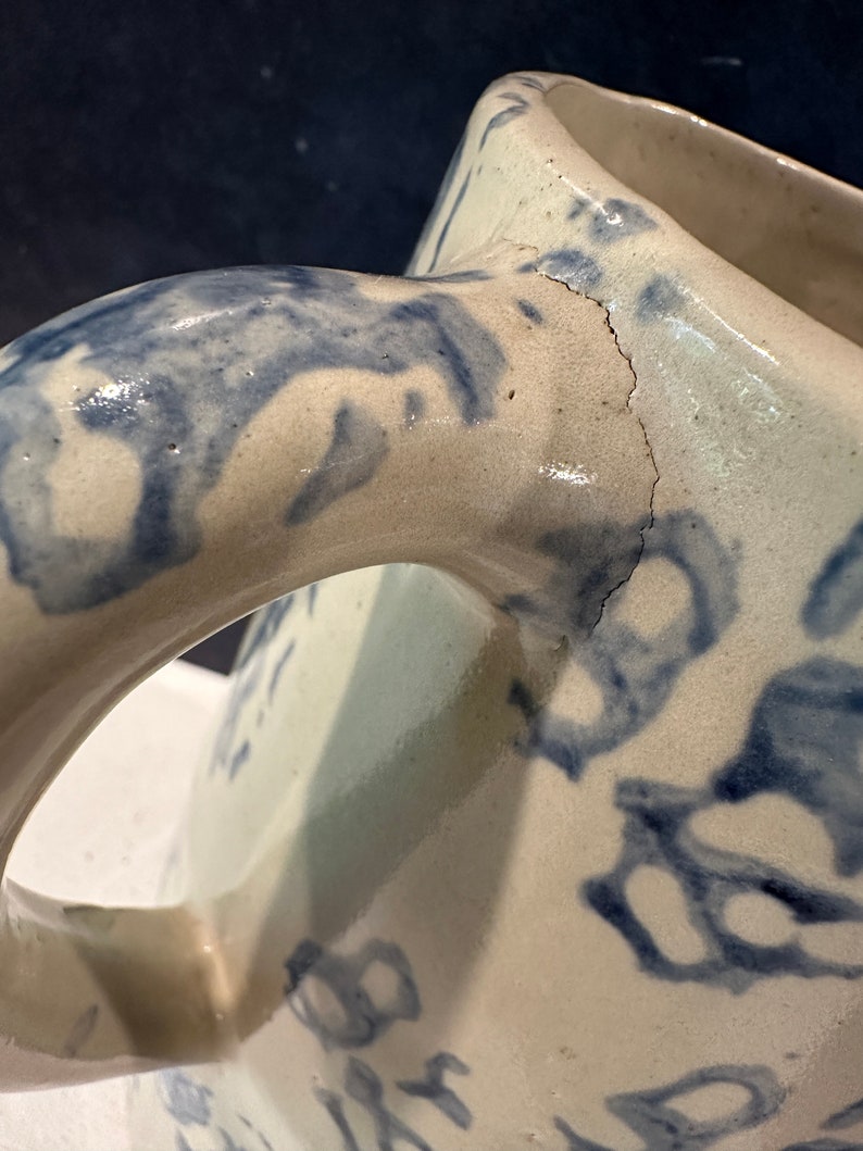 Op de afbeelding: Een witte keramische kan met een blauw bloemenmotief en een scheur in de handgreep.