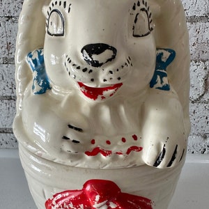May include: A vintage ceramic cookie jar shaped like a white bunny with blue bows and red accents. The bunny is sitting in a basket with a red bow.