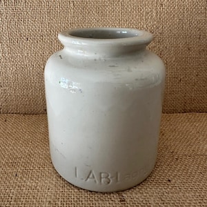 May include: A vintage, off-white ceramic jar with a rounded body and a slightly flared rim. The jar has a cylindrical shape and the word "LABIagre" is embossed on the lower portion. The jar is likely used for storage.