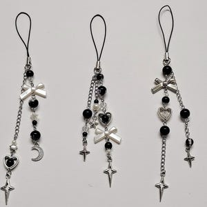 May include: Three black and silver phone charms with various charms. Each charm features black beads, a bow, a heart, a star, and a chain. One charm has a crescent moon. The charms hang from a black loop.