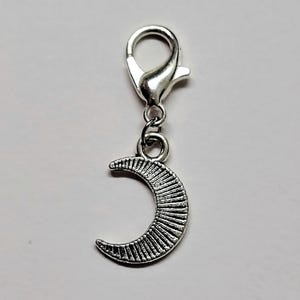 May include: Silver charm with a crescent moon design. The moon has a textured surface and is attached to a silver lobster clasp.