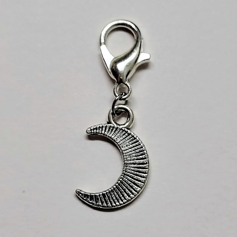 May include: Silver charm with a crescent moon design. The moon has a textured surface and is attached to a silver lobster clasp.
