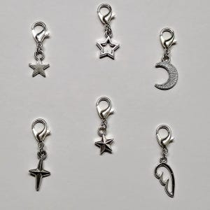 May include: Six silver charm clips with different designs: a star, a crescent moon, a star with a point, a star, a wing, and a star with a point.