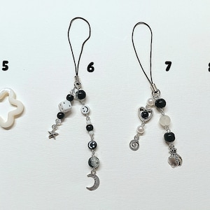 May include: Five black and white phone charms with different designs. The charms are made with beads, metal charms, and a white star. The charms are attached to black cords.