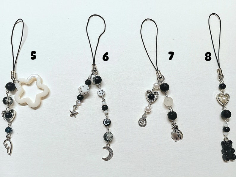 May include: Five black and white phone charms with different designs. The charms are made with beads, metal charms, and a white star. The charms are attached to black cords.