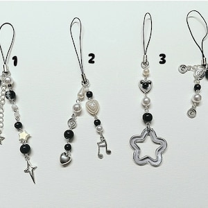 May include: Four black and white phone charms with different designs. The charms feature a crescent moon, stars, a heart, a music note, a star, and a swirl design.