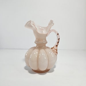 May include: A pale pink glass pitcher with a ruffled top and a twisted handle. The pitcher has a bulbous body with a textured surface.