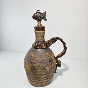 May include: A brown ceramic pitcher with a fish-shaped lid and a brown handle. The pitcher has a textured surface and a rustic look.