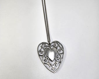 May include: A silver-toned pendant necklace featuring a heart-shaped design. The pendant has a filigree outer heart surrounding a solid heart. The necklace chain is a delicate silver color.