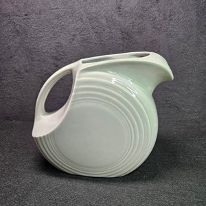 May include: A white ceramic pitcher with a unique, rounded design and a wide, flared spout. The pitcher has a smooth, glossy finish and a subtle, rippled pattern.