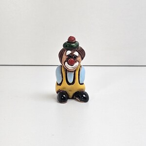 May include: A small ceramic clown figurine. The clown has brown hair, a red nose, and a white smile. It wears a yellow overall with blue sleeves and a green hat with a red pom-pom. The clown's shoes are black.