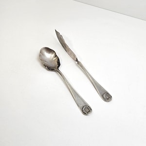 May include: A silver-toned metal spoon and a matching butter knife. Both utensils feature a scalloped shell-like design at the base of the handle. The spoon has a rounded bowl, while the knife has a flat, slightly curved blade edge.