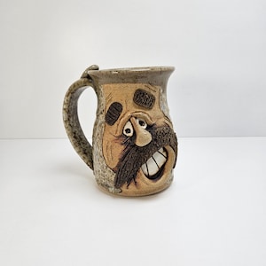May include: A whimsical, handmade ceramic mug with a sculpted face. The mug features a tan and brown color scheme, with a textured handle and a detailed, expressive face with a mustache and teeth. The mug is designed for beverages.