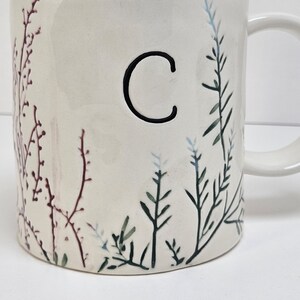 K&ouml;nnte beinhalten: Cremefarbene Keramiktasse mit einem schwarzen Buchstaben "C" und botanischen Illustrationen. Die Tasse hat einen Henkel und ist mit Zweigen in Gr&uuml;n-, Blau- und Burgundert&ouml;nen verziert. Das Design ist auf die Au&szlig;enseite der Tasse gemalt.