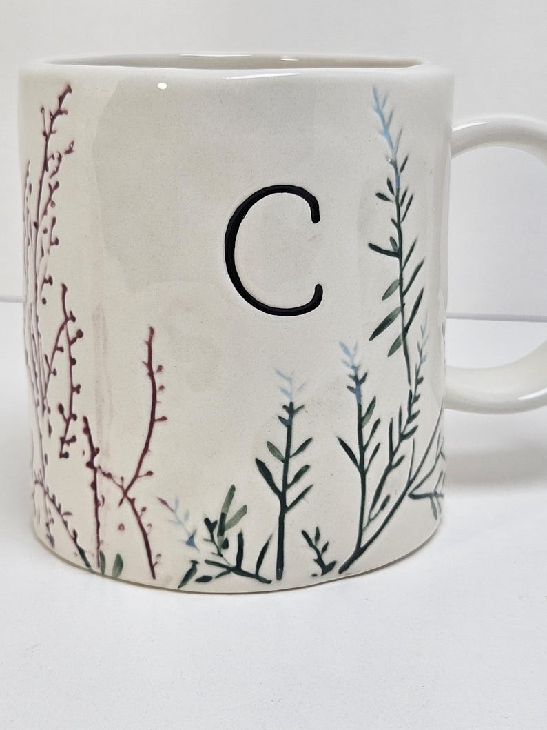 K&ouml;nnte beinhalten: Cremefarbene Keramiktasse mit einem schwarzen Buchstaben "C" und botanischen Illustrationen. Die Tasse hat einen Henkel und ist mit Zweigen in Gr&uuml;n-, Blau- und Burgundert&ouml;nen verziert. Das Design ist auf die Au&szlig;enseite der Tasse gemalt.
