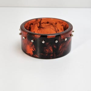 May include: A wide, round bangle bracelet with a marbled pattern in shades of orange and black. The bracelet is adorned with small, silver-colored studs and clear, sparkling accents. The jewelry piece is set against a white background.