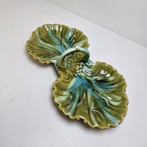 May include: A ceramic serving dish with a leaf-like design. The dish is divided into two sections and features a handle. The colors are shades of green and blue with a glossy finish. The dish is likely used for serving snacks or appetizers.